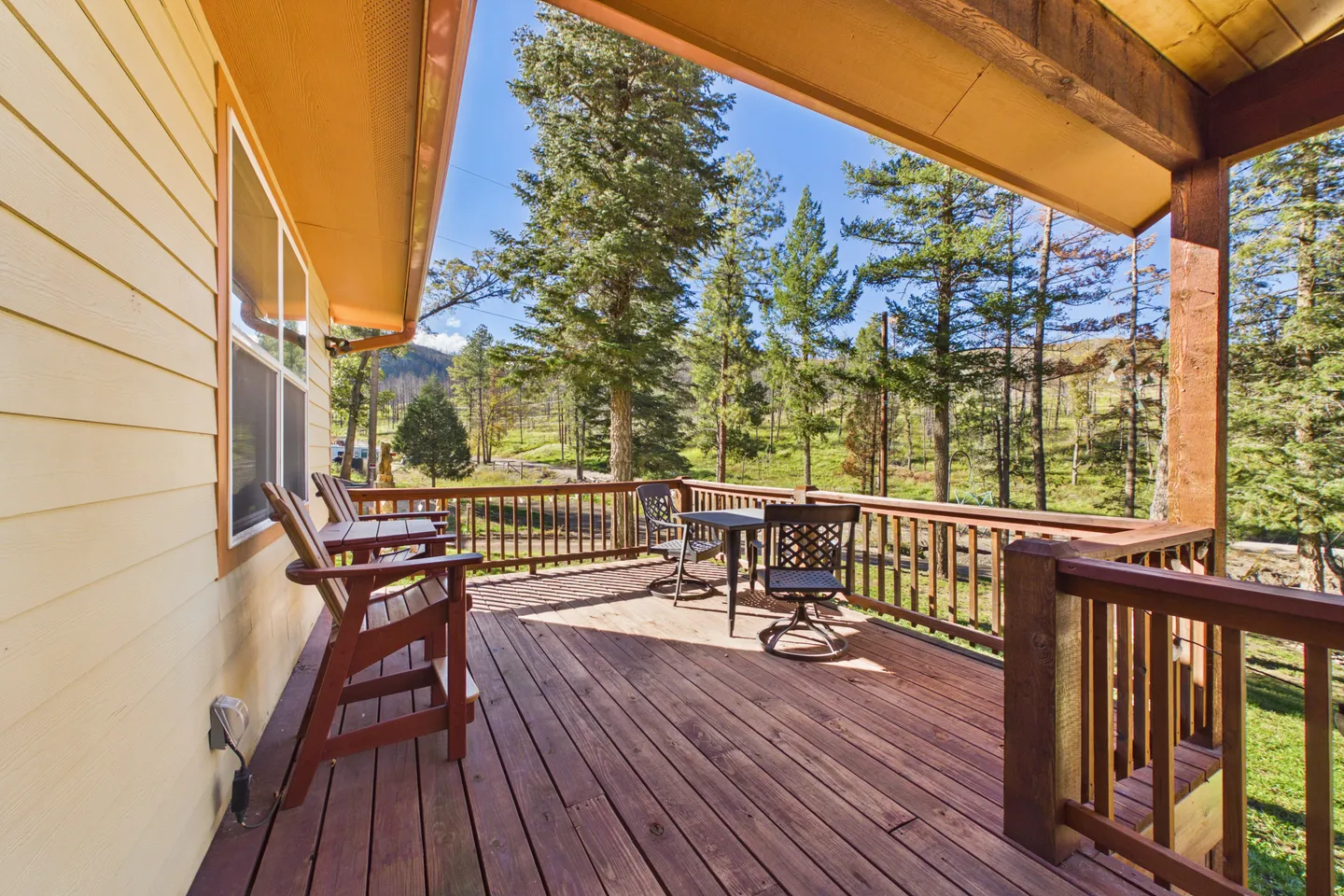 A wooden deck with railing, outdoor seating, and views of tall evergreen trees under a bright blue sky.