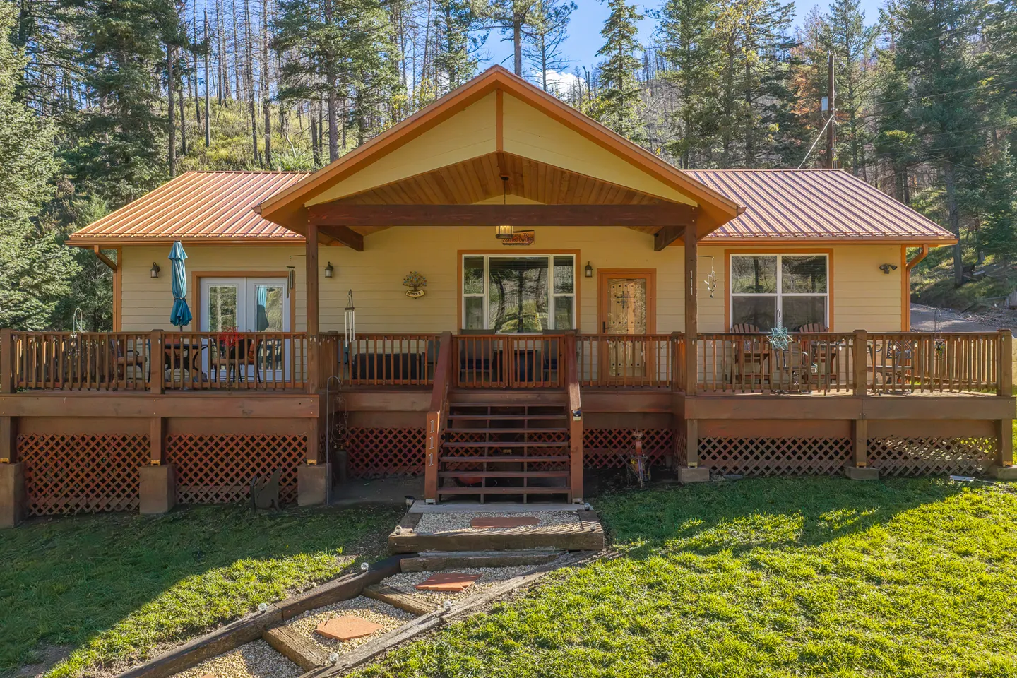 Exterior front view of a single-story home with a gabled metal roof, elevated wooden deck, and front steps leading to the lawn.