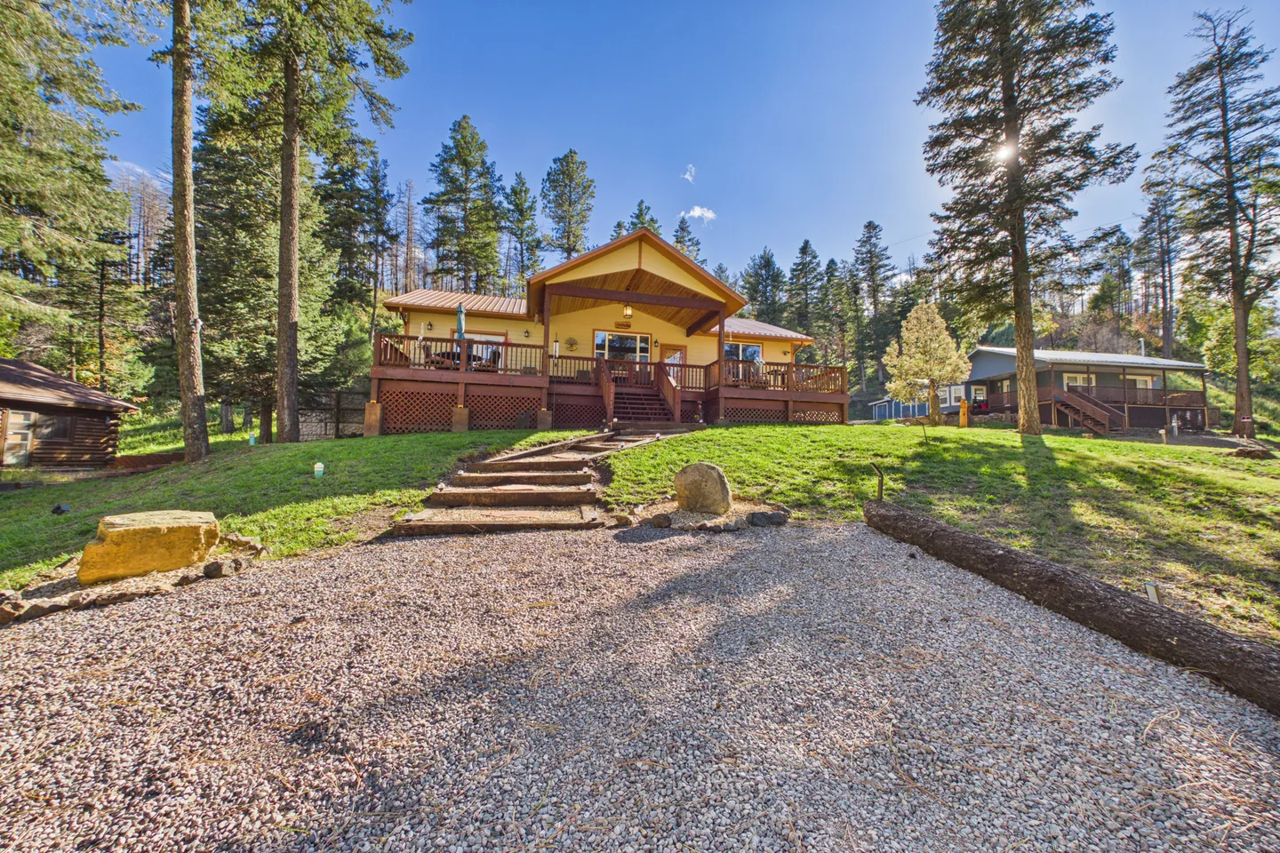 Exterior view of a yellow home with an elevated front porch and wooden steps leading down to a gravel driveway, framed by tall pine trees.
