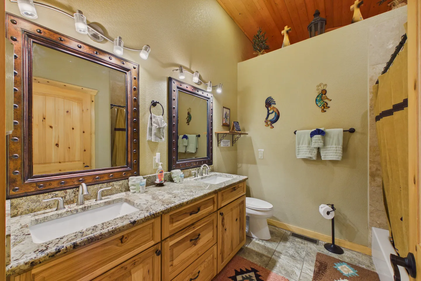 Bathroom with a double vanity featuring rustic wood cabinets, stone-look countertops, framed mirrors, and a toilet next to a shower area.