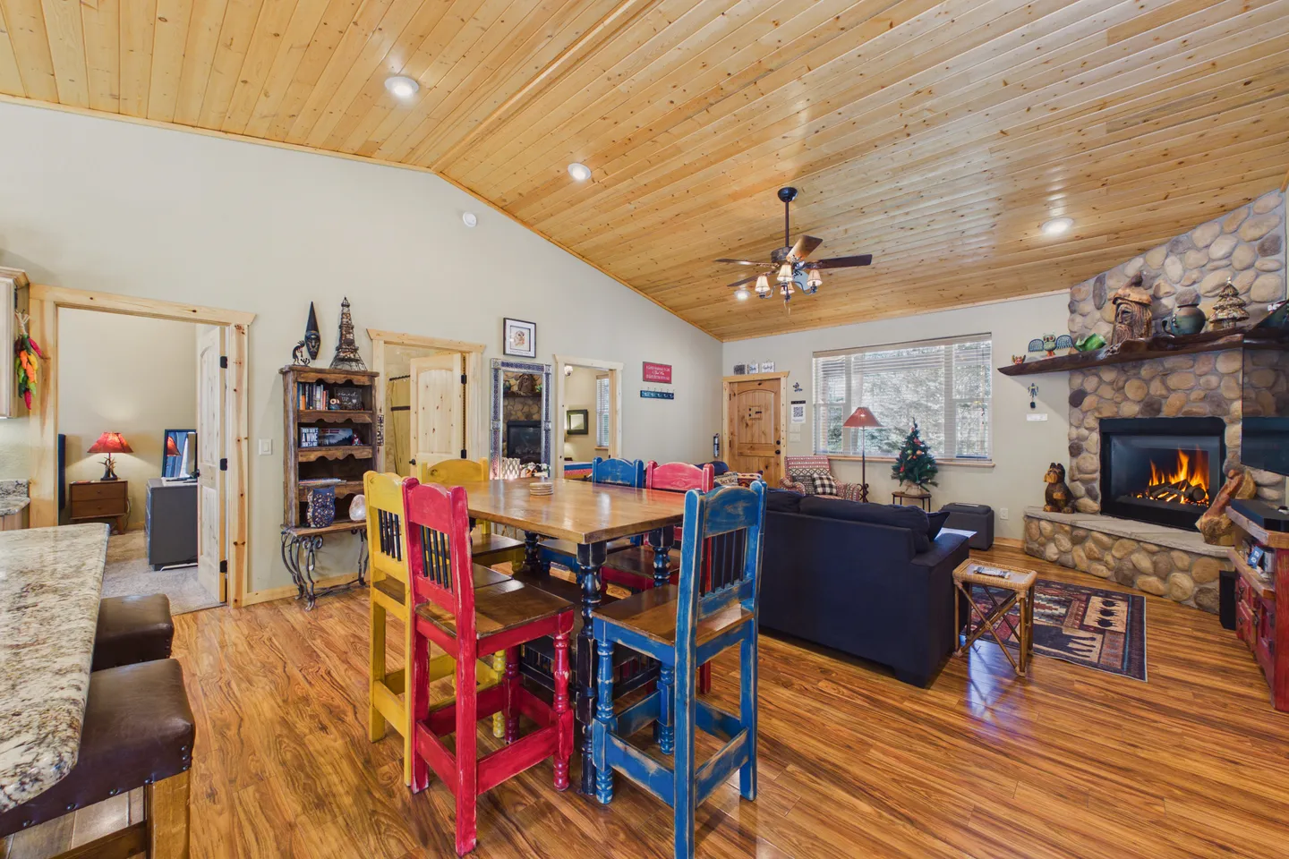 Open-concept dining and living room featuring vaulted wood-paneled ceiling, colorful high-top chairs at a dining table, and a stone corner fireplace.