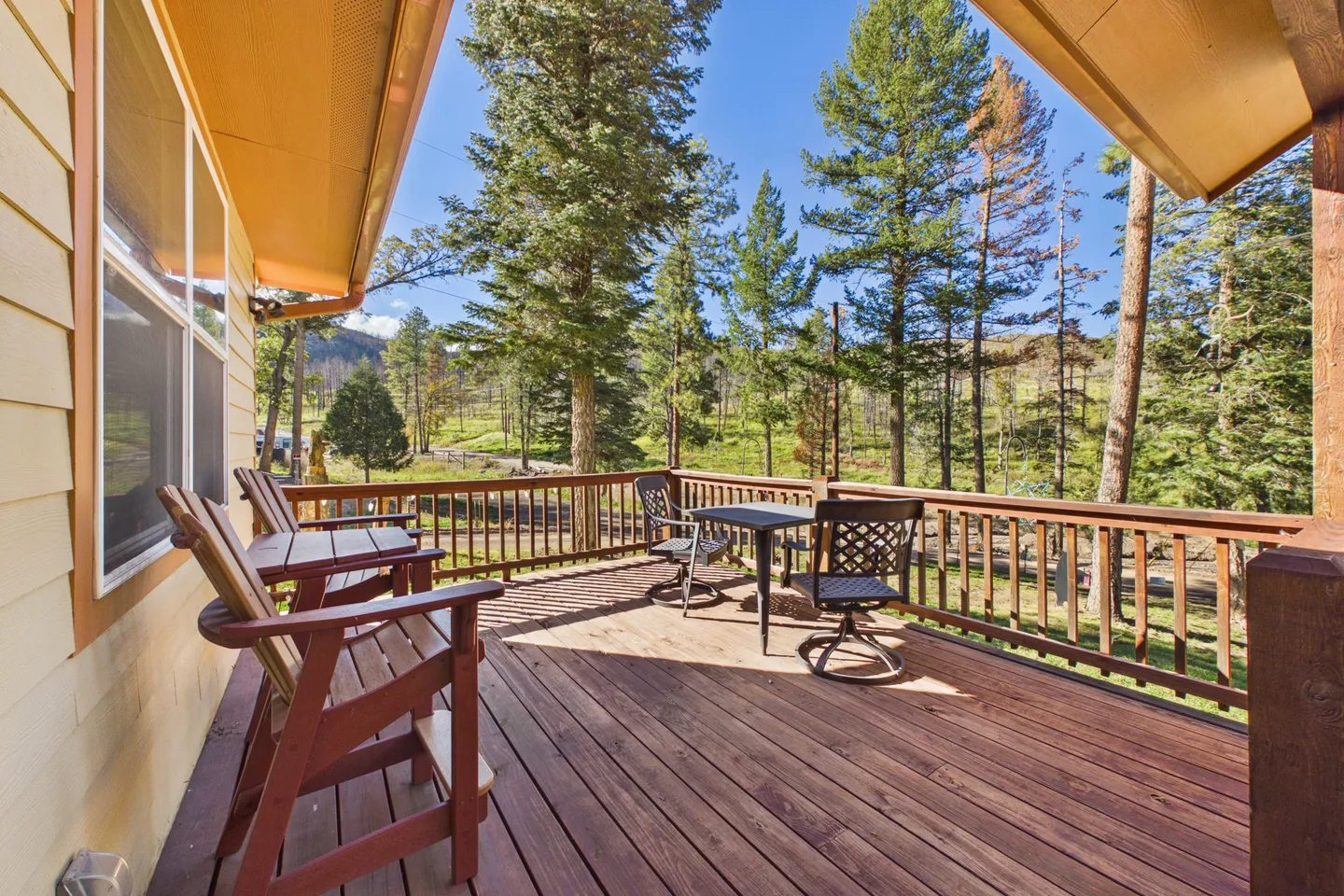Elevated wooden deck with outdoor chairs and table overlooking tall pine trees and a grassy hillside under a clear blue sky.