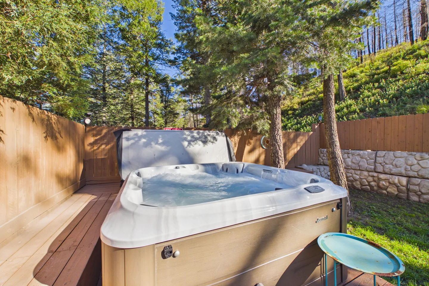 Outdoor hot tub on a wooden deck surrounded by trees and a wooden fence.