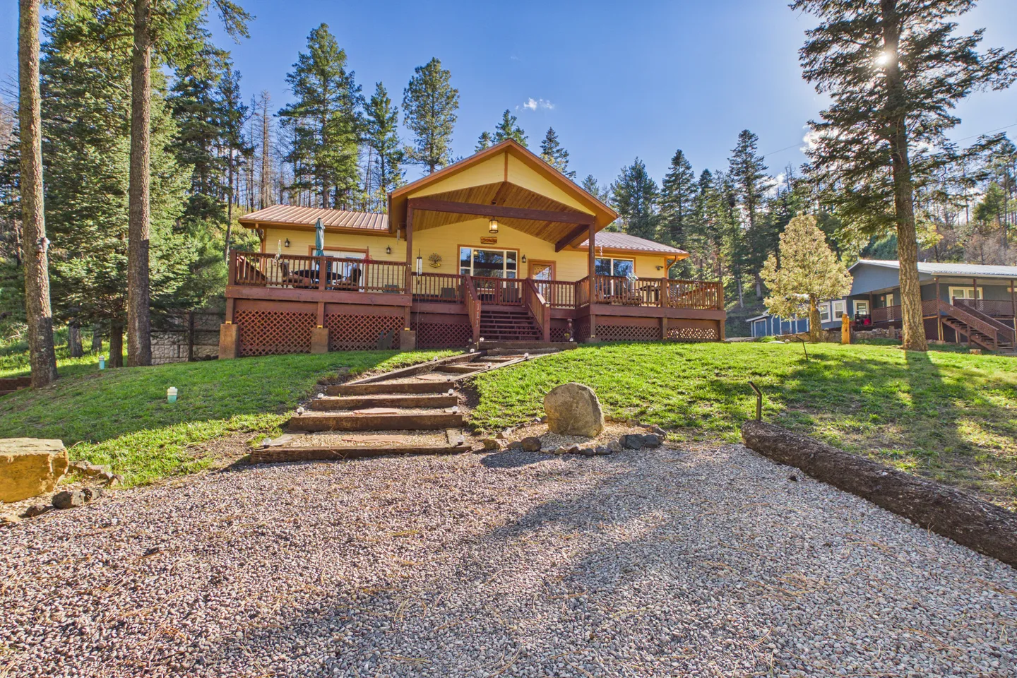Exterior view of a yellow single-story home with a large raised wooden deck, framed by pine trees under a clear blue sky.