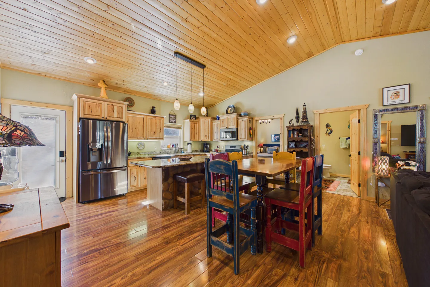 Open-concept kitchen and dining area with wood-paneled vaulted ceiling, stainless steel refrigerator, and rustic wooden dining set.