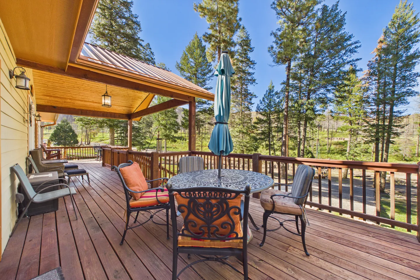 Spacious wooden outdoor deck featuring a patio dining table with an umbrella, chairs with cushions, and a view of pine trees under a clear blue sky.