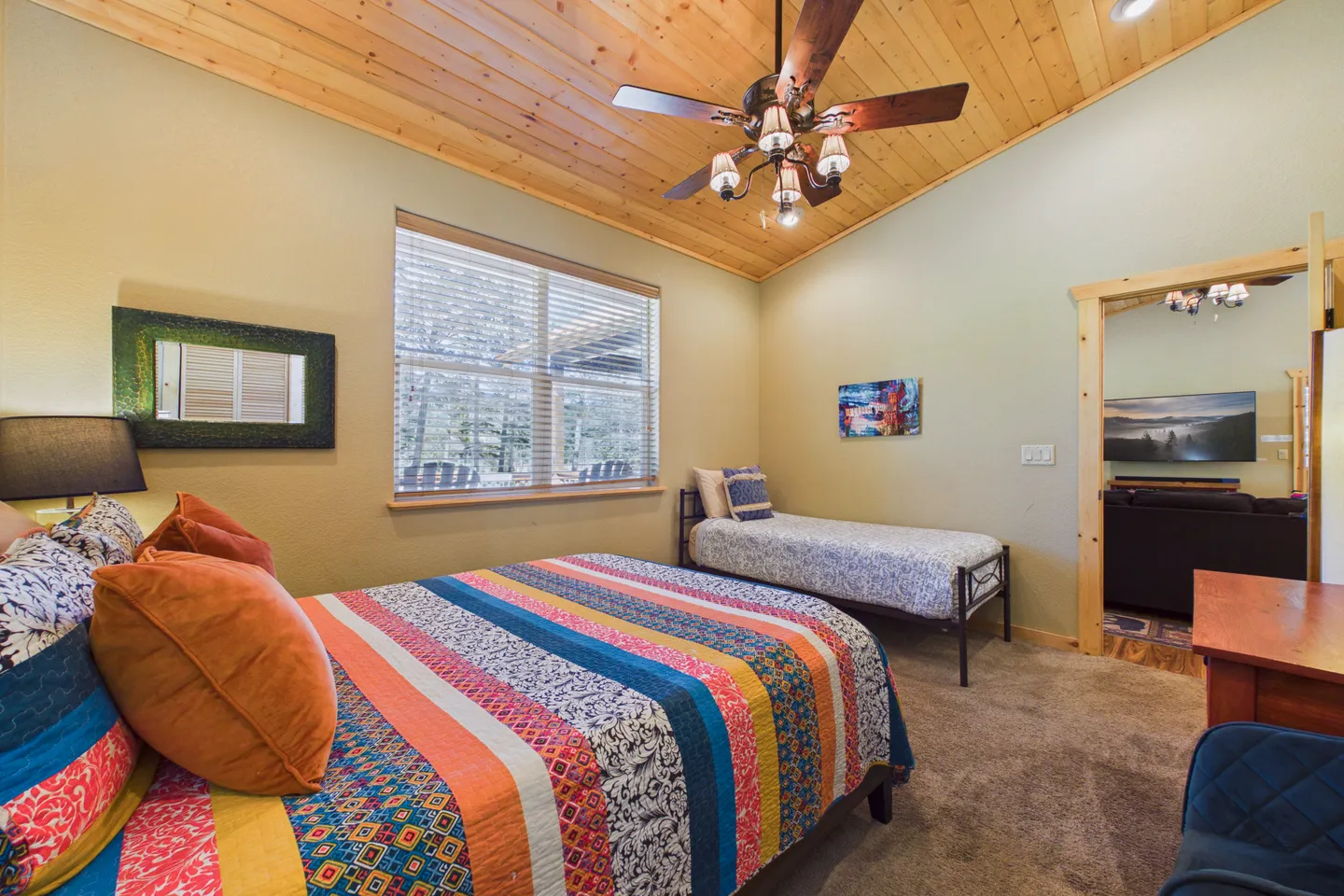 Bedroom with wood-plank ceiling, ceiling fan, a double bed with a colorful striped quilt, a single bed along the wall, and carpeted flooring.
