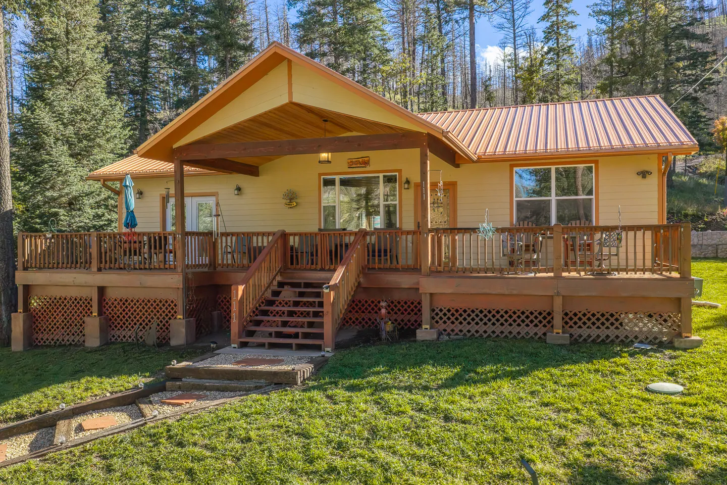 Exterior front view of a single-story yellow house with a covered wooden porch, metal roof, and surrounding pine forest.
