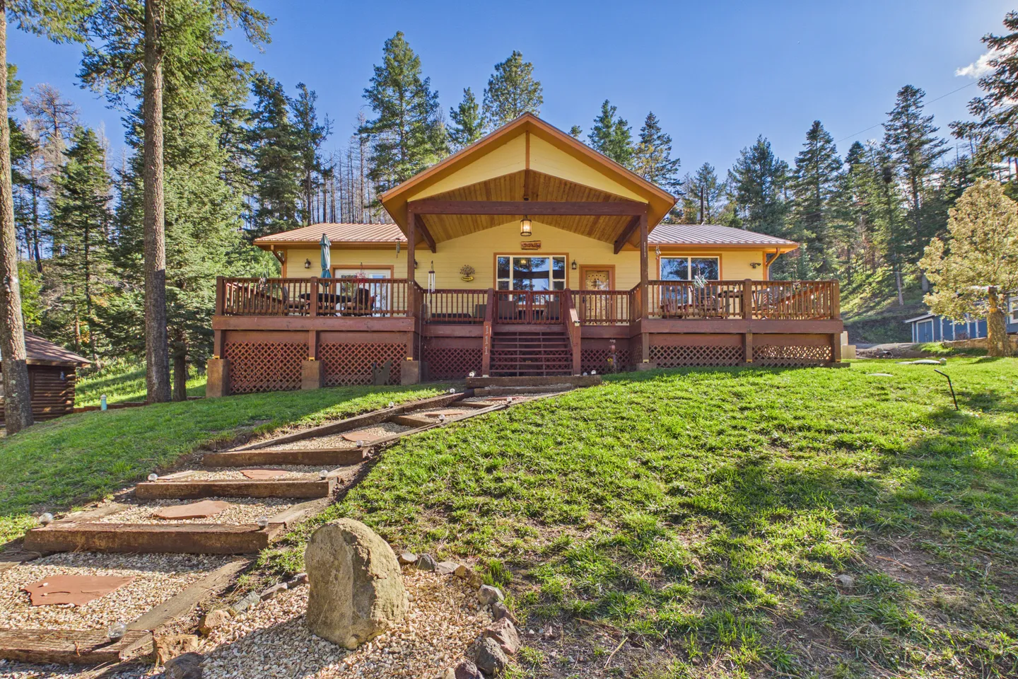 Exterior view of a yellow cabin with a large elevated wooden deck set on a grassy hill surrounded by pine trees under a clear blue sky.