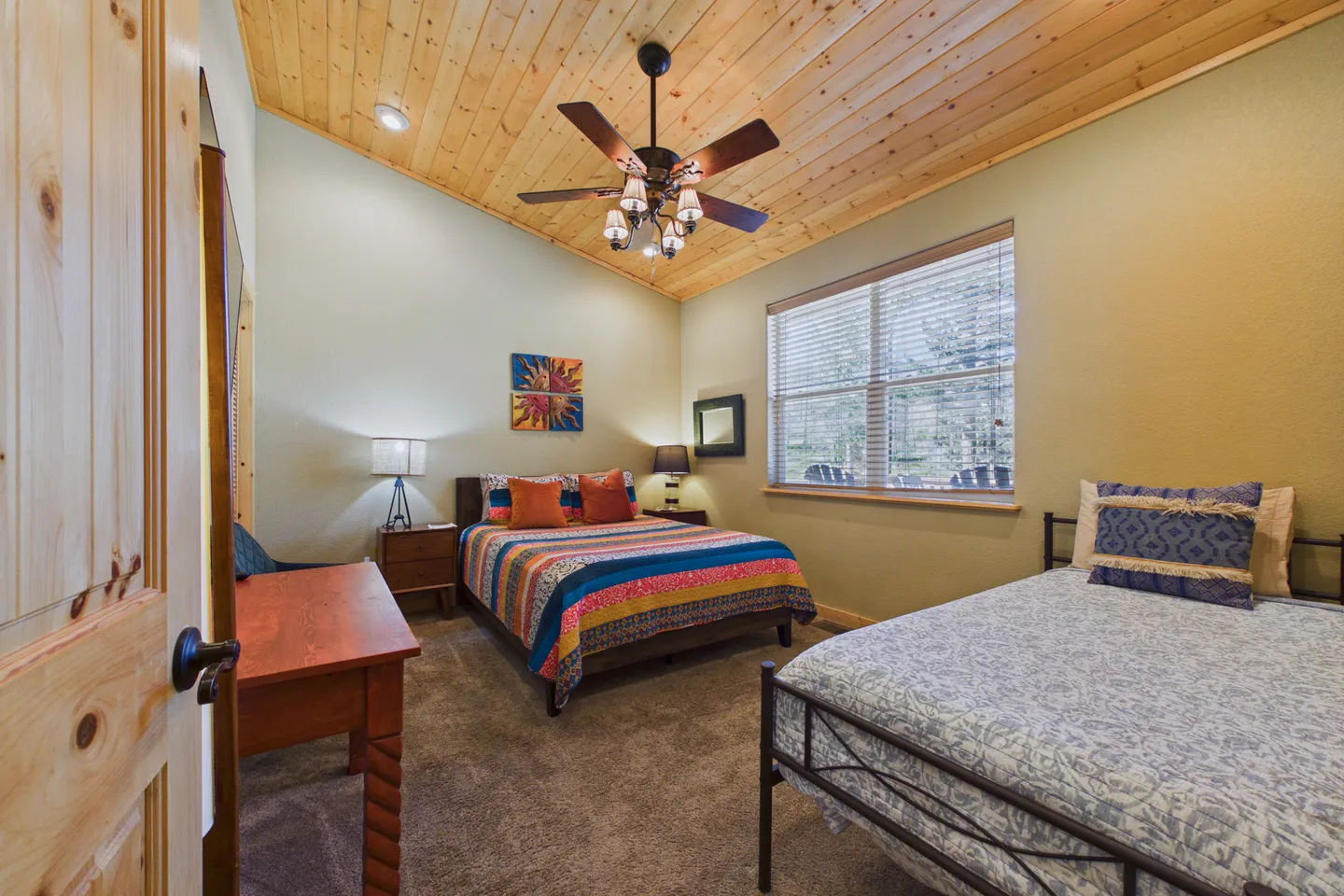 Bedroom with vaulted wood-paneled ceiling, two beds, a desk, and a window with blinds.