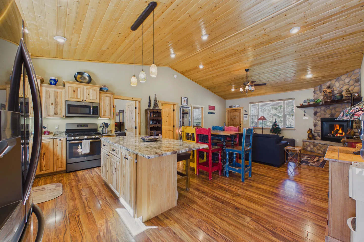Open-concept cabin interior featuring a kitchen island with granite countertop, colorful dining chairs, vaulted wood-paneled ceiling, and a stone fireplace in the living area.