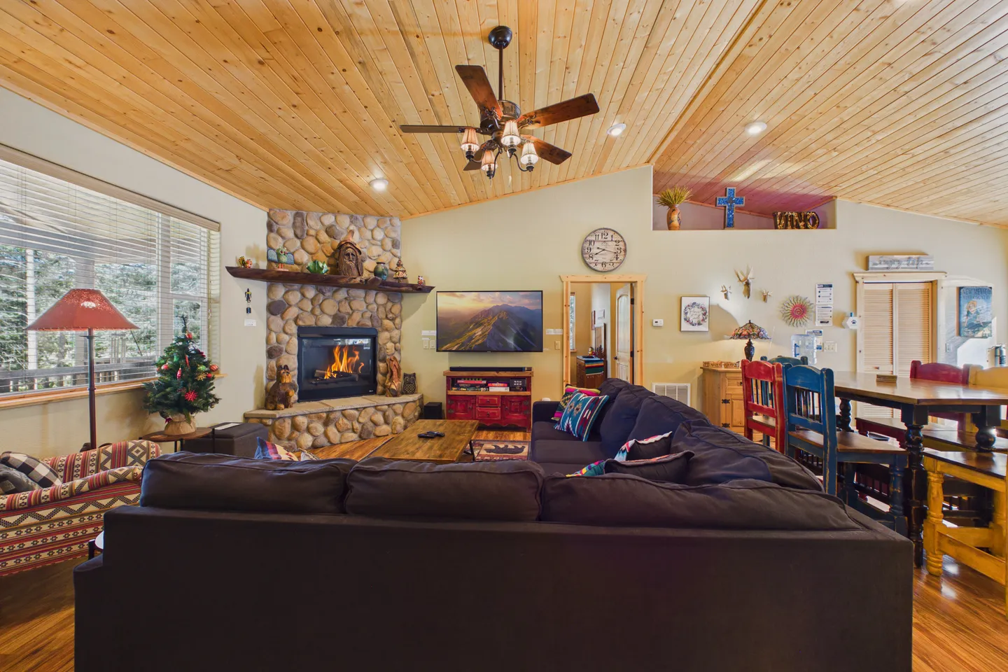 Living room with wood-paneled angled ceiling, corner stone fireplace with fire burning, TV mounted on the wall, and dark sectional sofa.