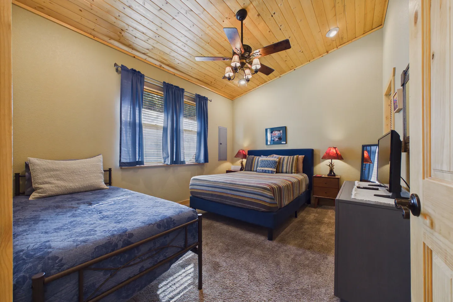 Bedroom with vaulted wood-plank ceiling, ceiling fan, carpet, two beds, and blue curtains.
