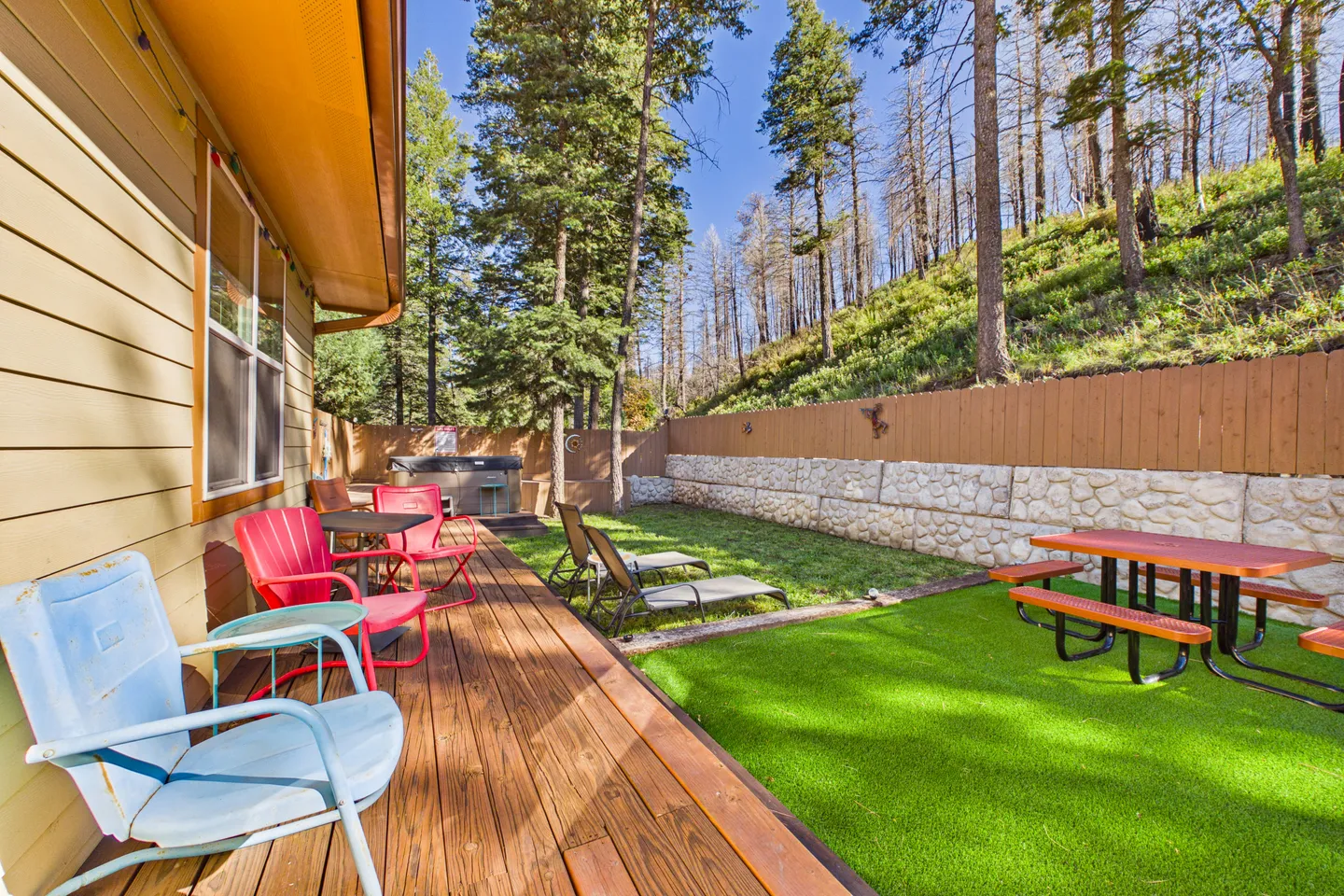 Fenced backyard featuring a wooden deck with outdoor chairs, an artificial turf area with a picnic table, lounge chairs, a hot tub, and a hillside forest in the background.