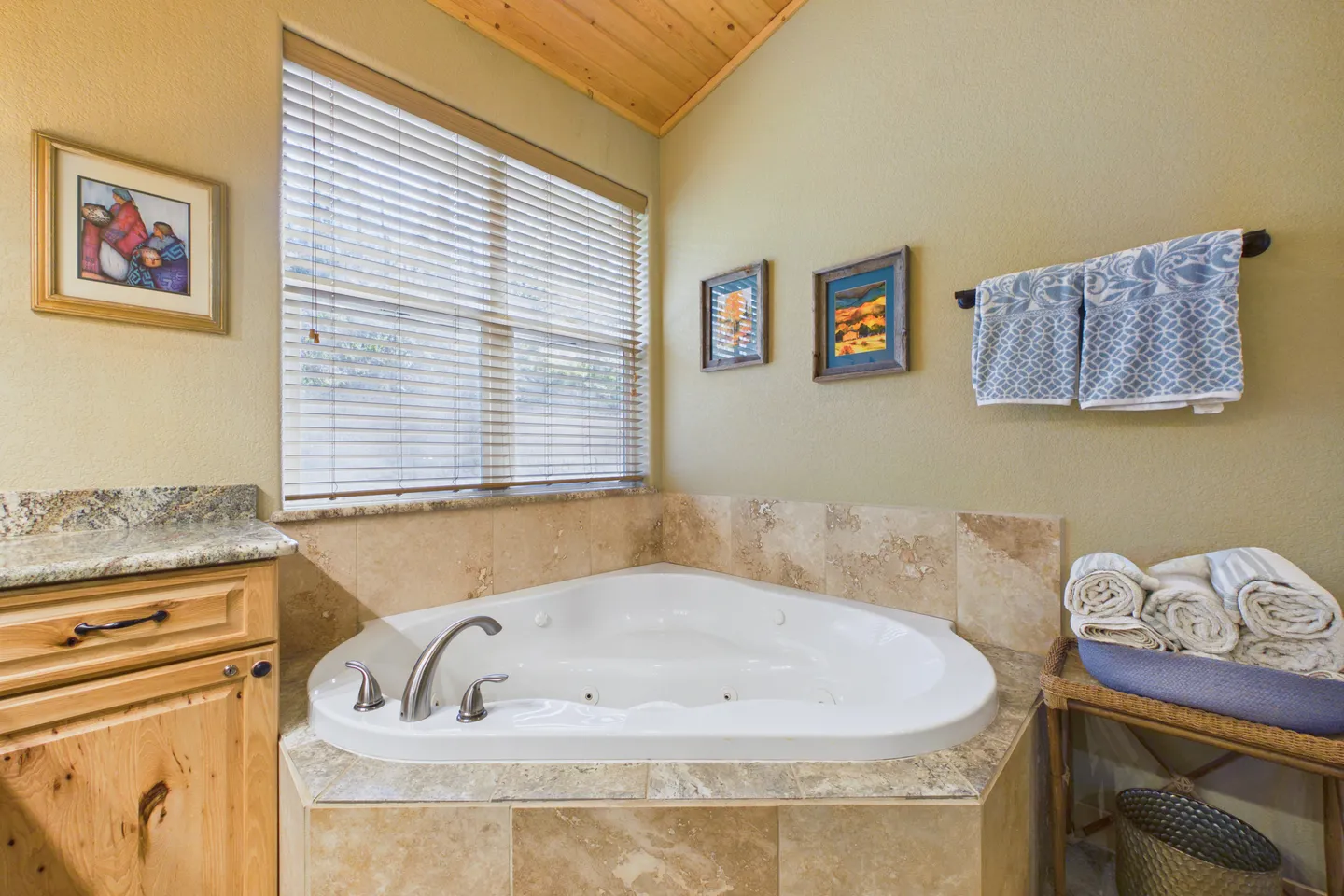A corner jetted soaking tub set into tiled surround beneath a window with blinds in a bathroom.