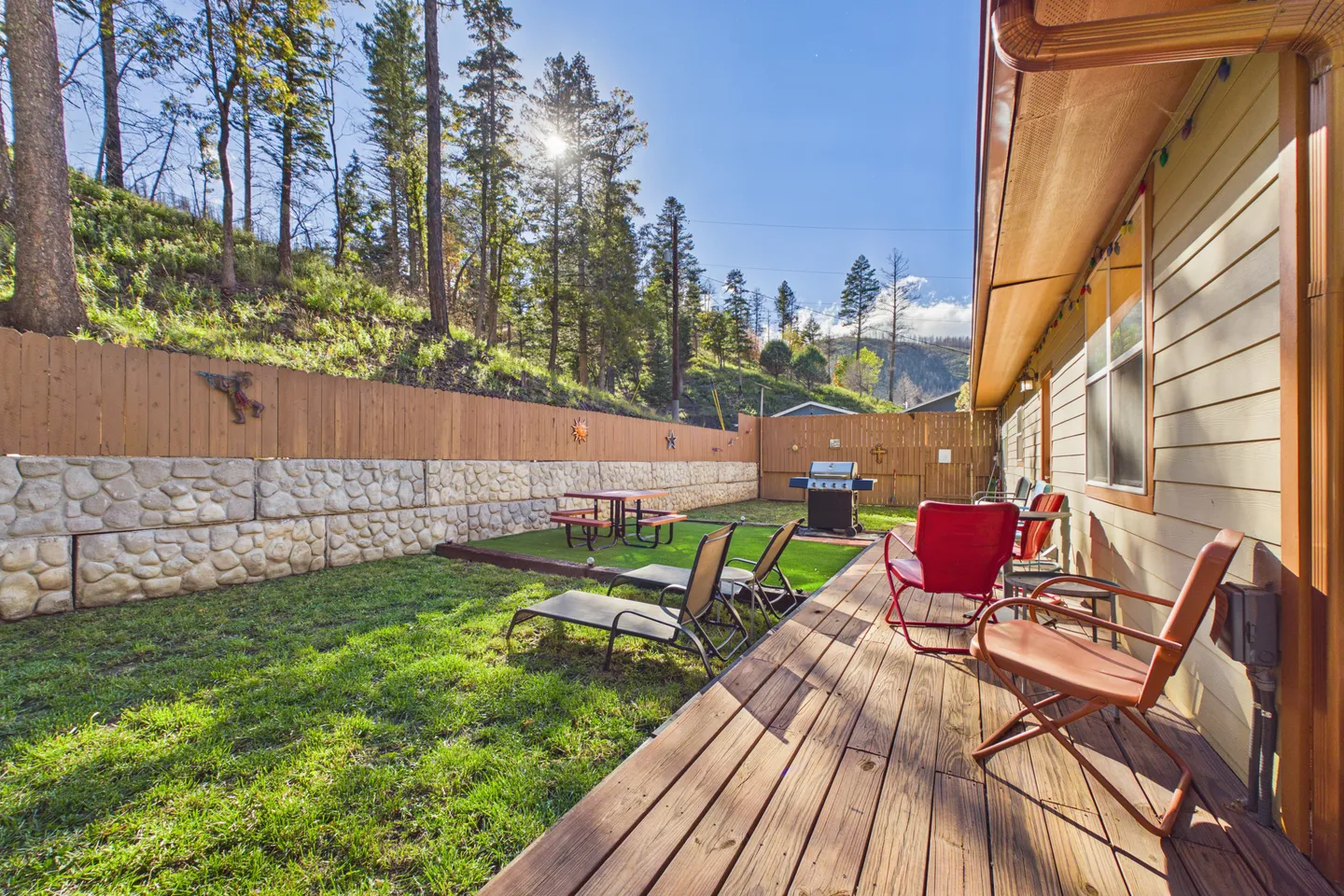Fenced backyard featuring a wooden deck with chairs, lawn chairs on grass, a barbecue grill, picnic table, and a wooded hillside background.