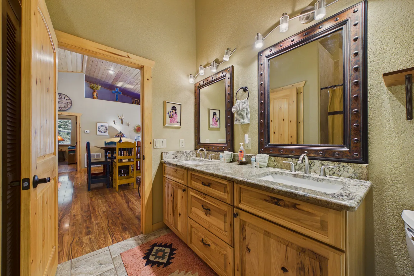 Bathroom with a double vanity featuring knotty pine cabinets, stone countertops, and two rustic framed mirrors, looking out into a living area.