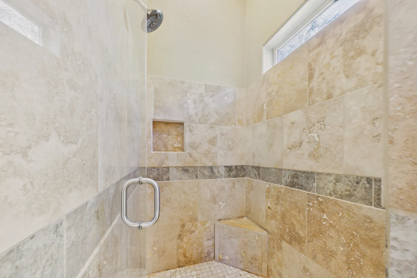 Walk-in tiled shower with a glass door, corner bench, recessed shelf, and small high window.