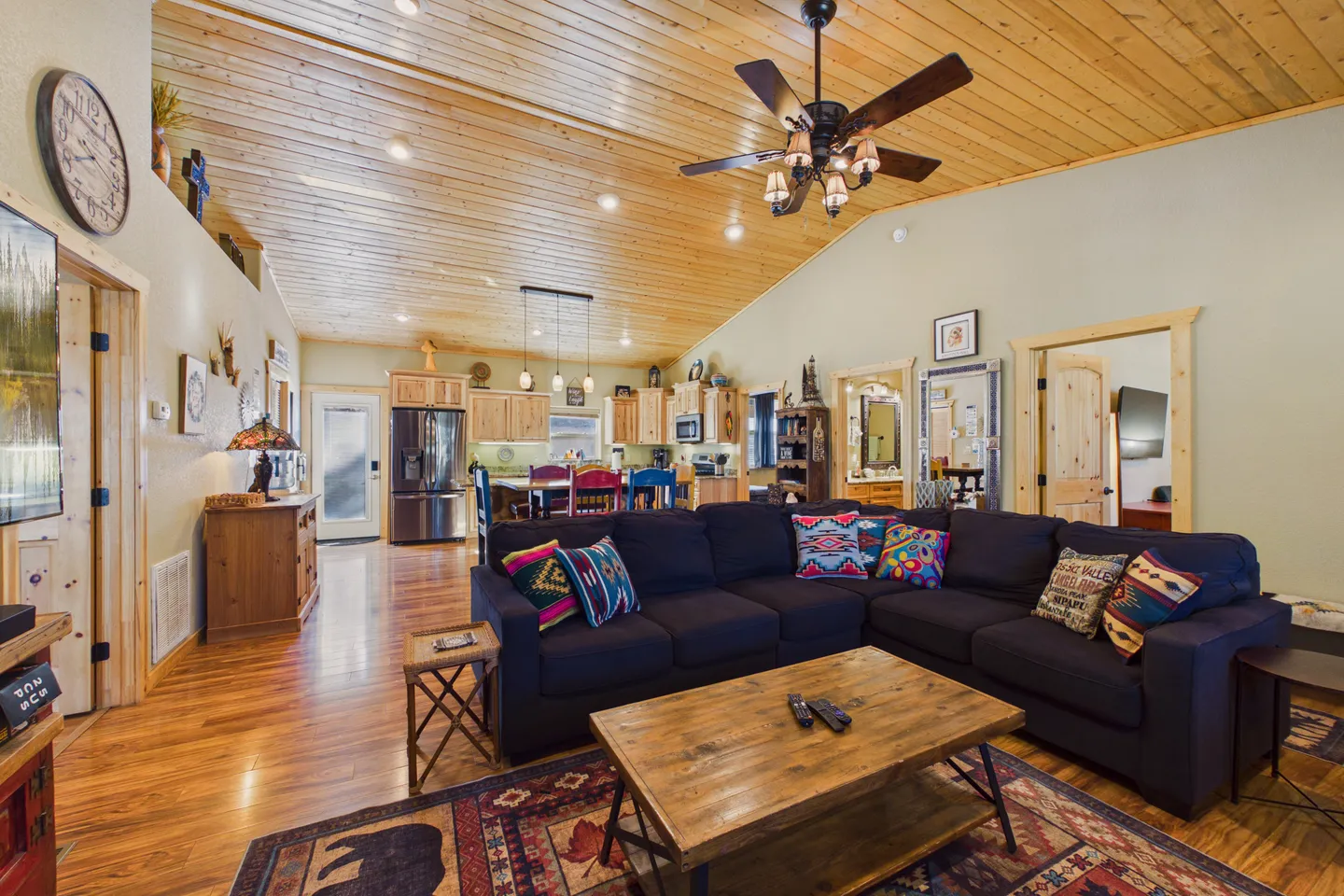 Open concept living room and kitchen featuring a wood-plank vaulted ceiling, a large dark sectional sofa, and wood flooring.