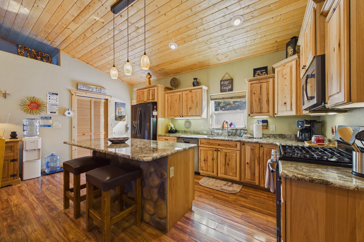 Kitchen with wood-plank sloped ceiling, knotty wood cabinets, granite countertops, and an island with stools.