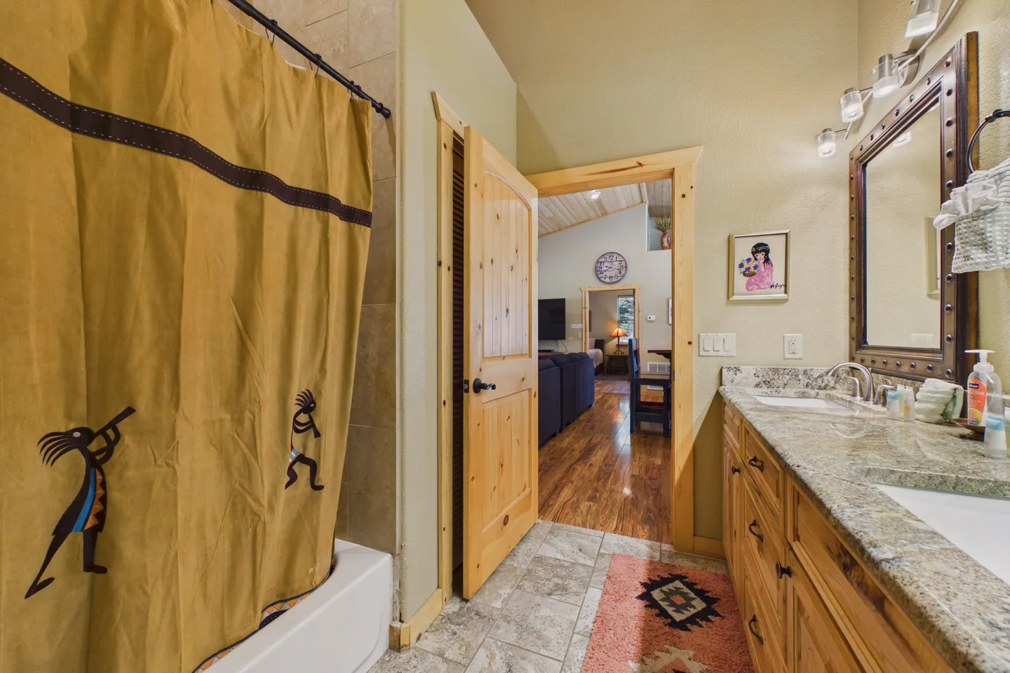 Bathroom with a shower curtain featuring Kokopelli figures, a double-sink stone vanity, and an open wood door looking into a living room.