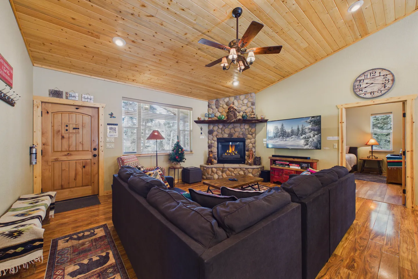Living room featuring wood-paneled angled ceiling, corner stone fireplace with burning fire, wall-mounted television, and a large sectional sofa.