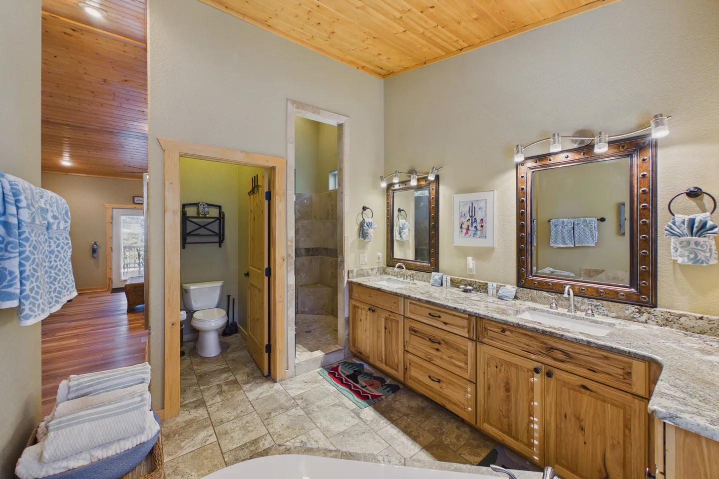 Bathroom with a double-sink wood vanity, tiled floor, wood-paneled ceiling, open tiled shower, and a private toilet room.