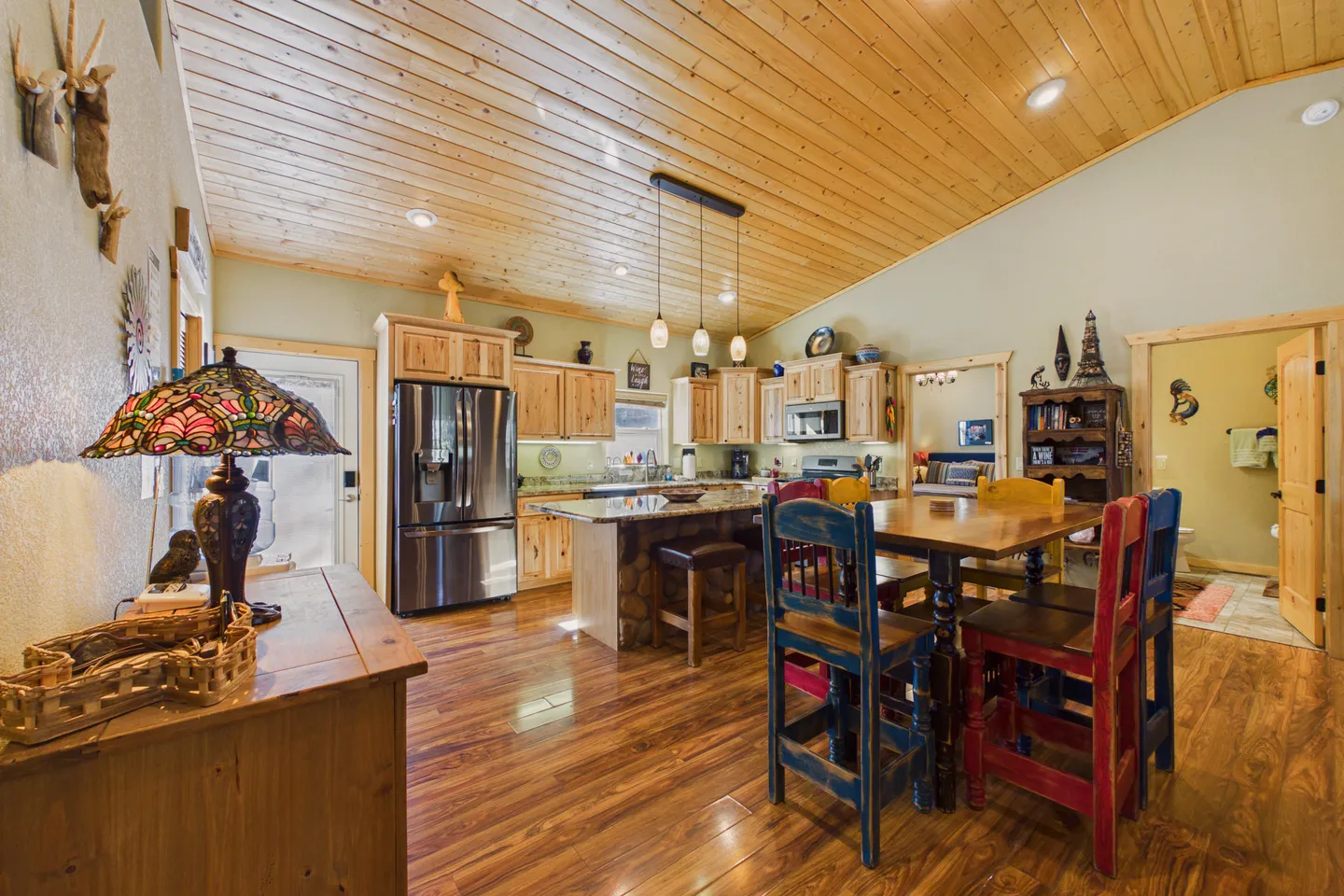 Open-concept kitchen and dining area with wood-paneled angled ceiling, stainless steel refrigerator, island, and dining table with colorful chairs.