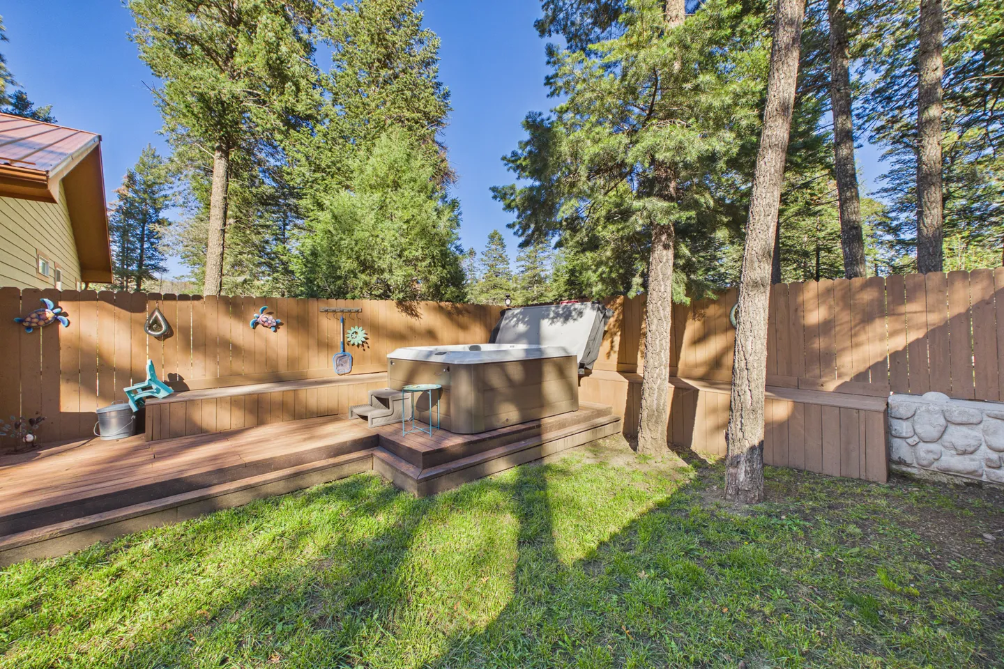 Outdoor hot tub set within a multi-level wooden deck in a grassy backyard enclosed by a tall wooden fence and pine trees.