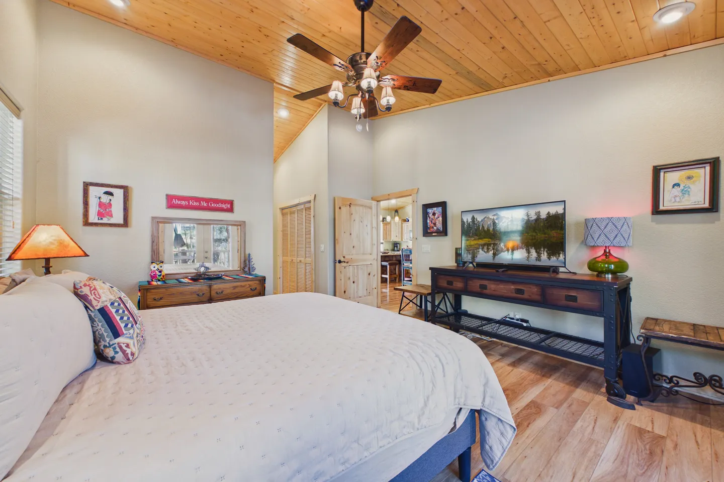 Bedroom with a vaulted wood-plank ceiling, ceiling fan, large bed, wooden dresser, and flat-screen TV on a rustic stand.