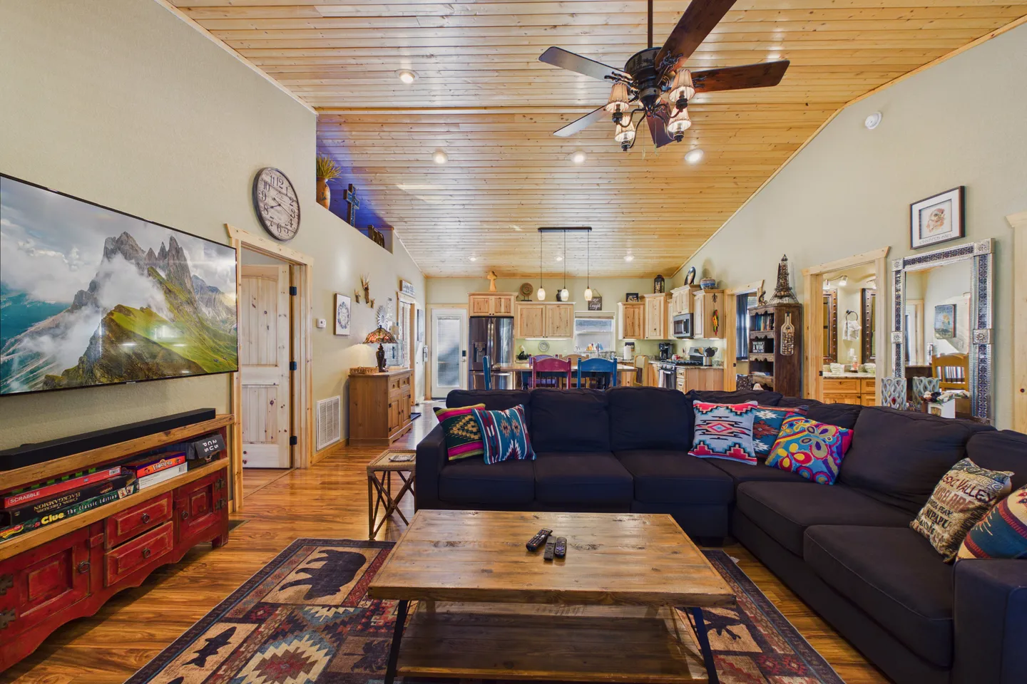 Open-concept living room with a large sectional sofa, wood coffee table, wall-mounted TV, and wood-paneled ceiling, looking toward the kitchen.