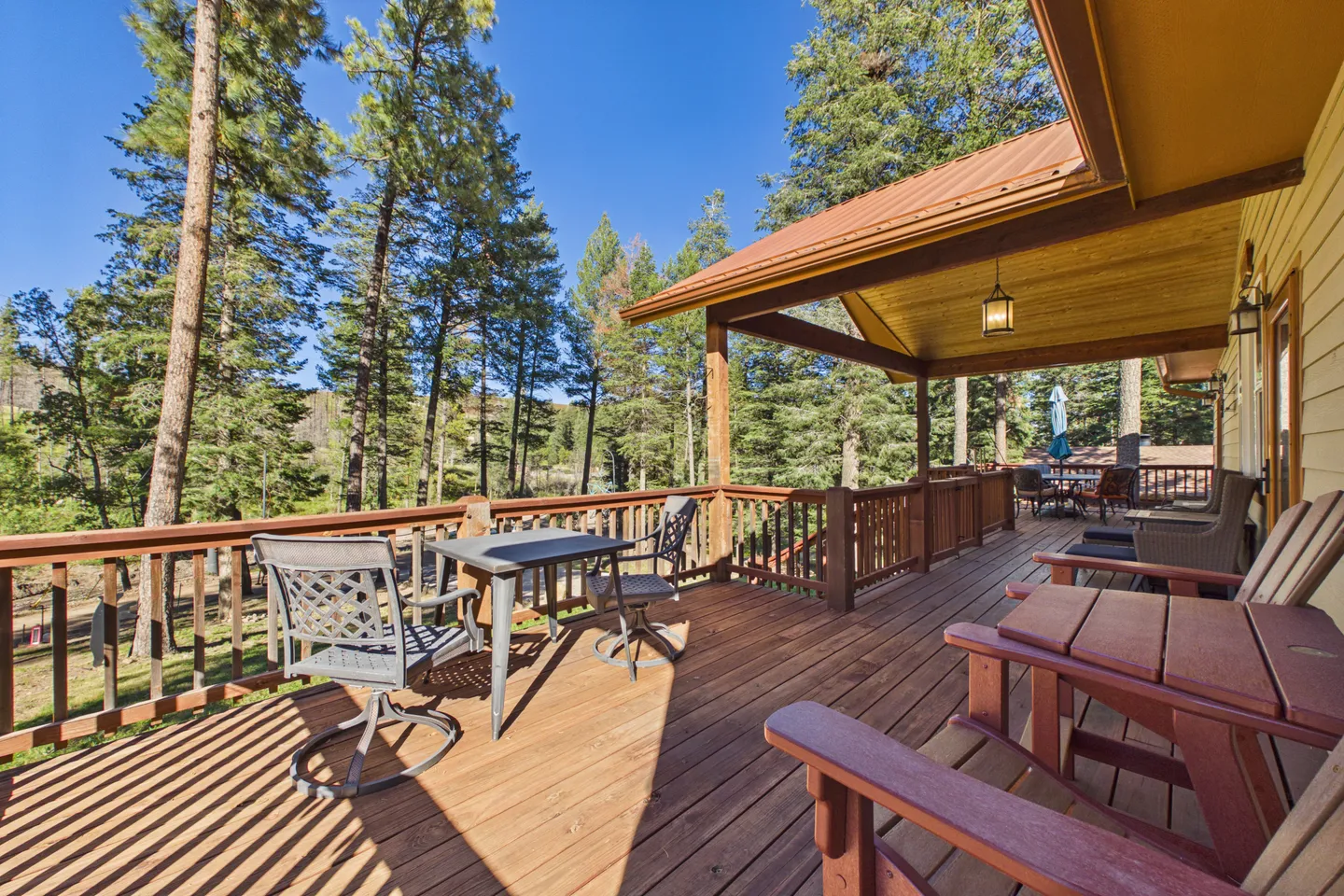 Spacious elevated wooden deck with patio chairs, table, covered porch area, and views of surrounding pine trees under a clear blue sky.