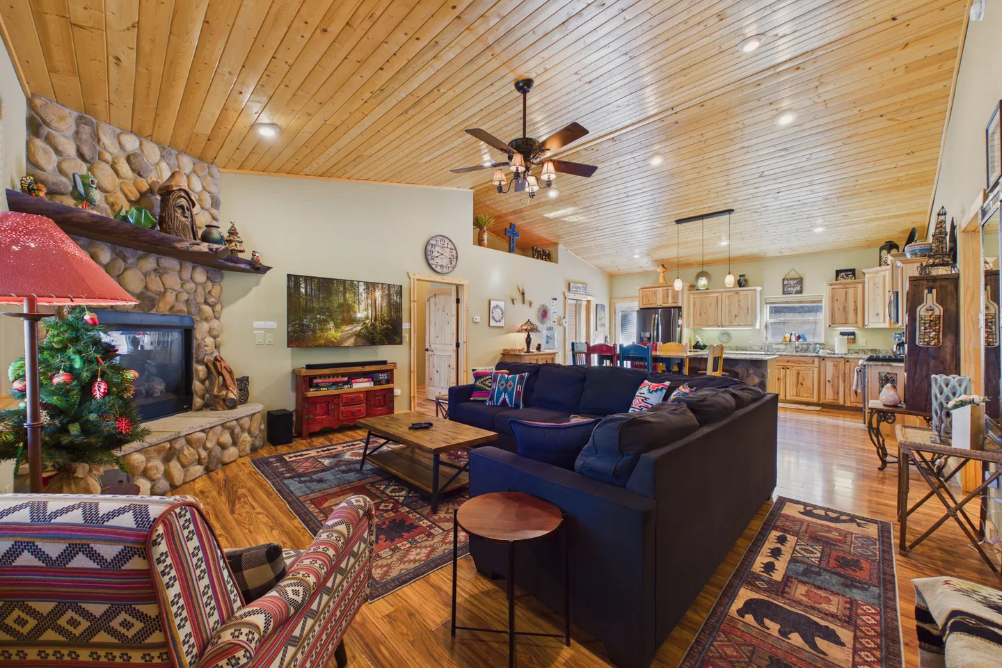 Open-concept living room and kitchen featuring a stone fireplace, wood-paneled ceiling, and dark sectional sofa.
