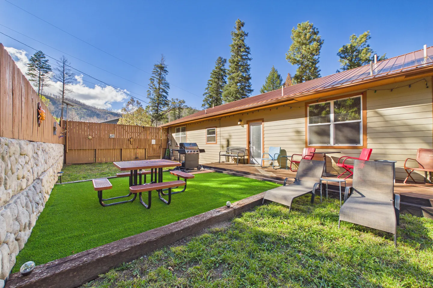 Fenced backyard featuring an artificial turf area with a picnic table, lounge chairs, a wooden patio with seating and a barbecue grill, and mountain views.