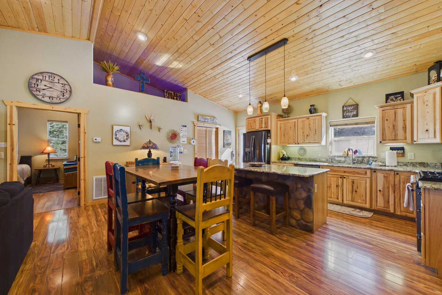 Open concept kitchen and dining space with wood-paneled ceiling, rustic wooden cabinets, kitchen island, and dining table with colorful chairs.