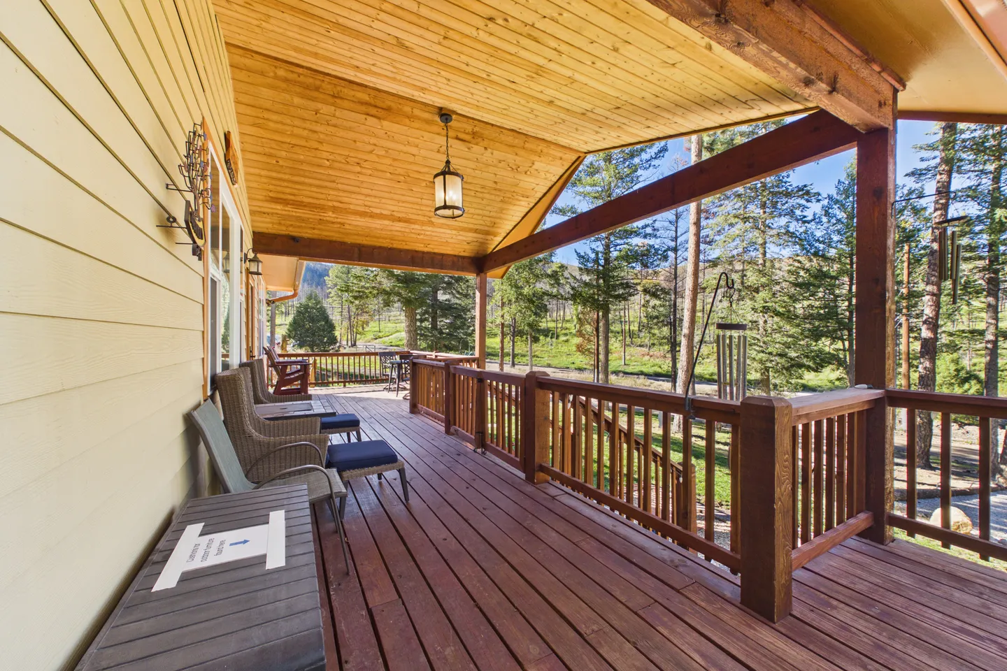 Covered wooden porch with seating and railings overlooking a wooded outdoor area under sunny skies.