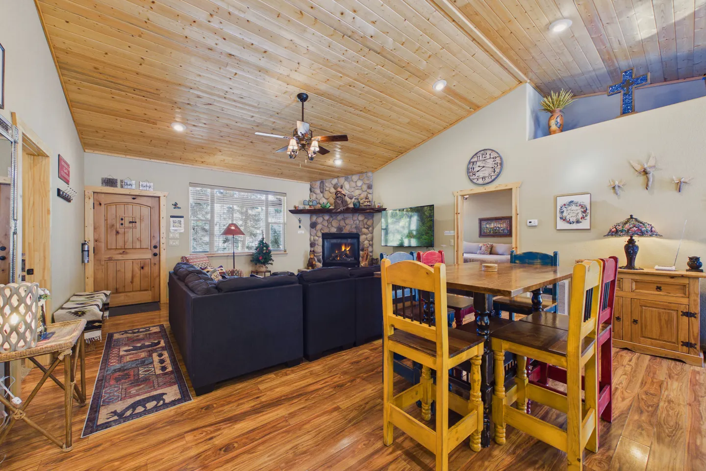 Open living and dining area featuring vaulted wood-paneled ceilings, a stone fireplace, sectional sofa, and a dining table with colorful chairs.