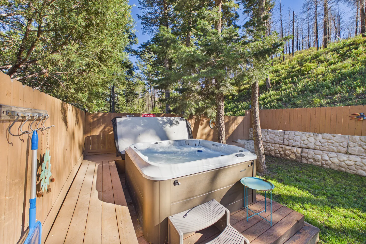 Outdoor hot tub on a wooden deck surrounded by a wooden privacy fence, trees, and a grassy hillside.
