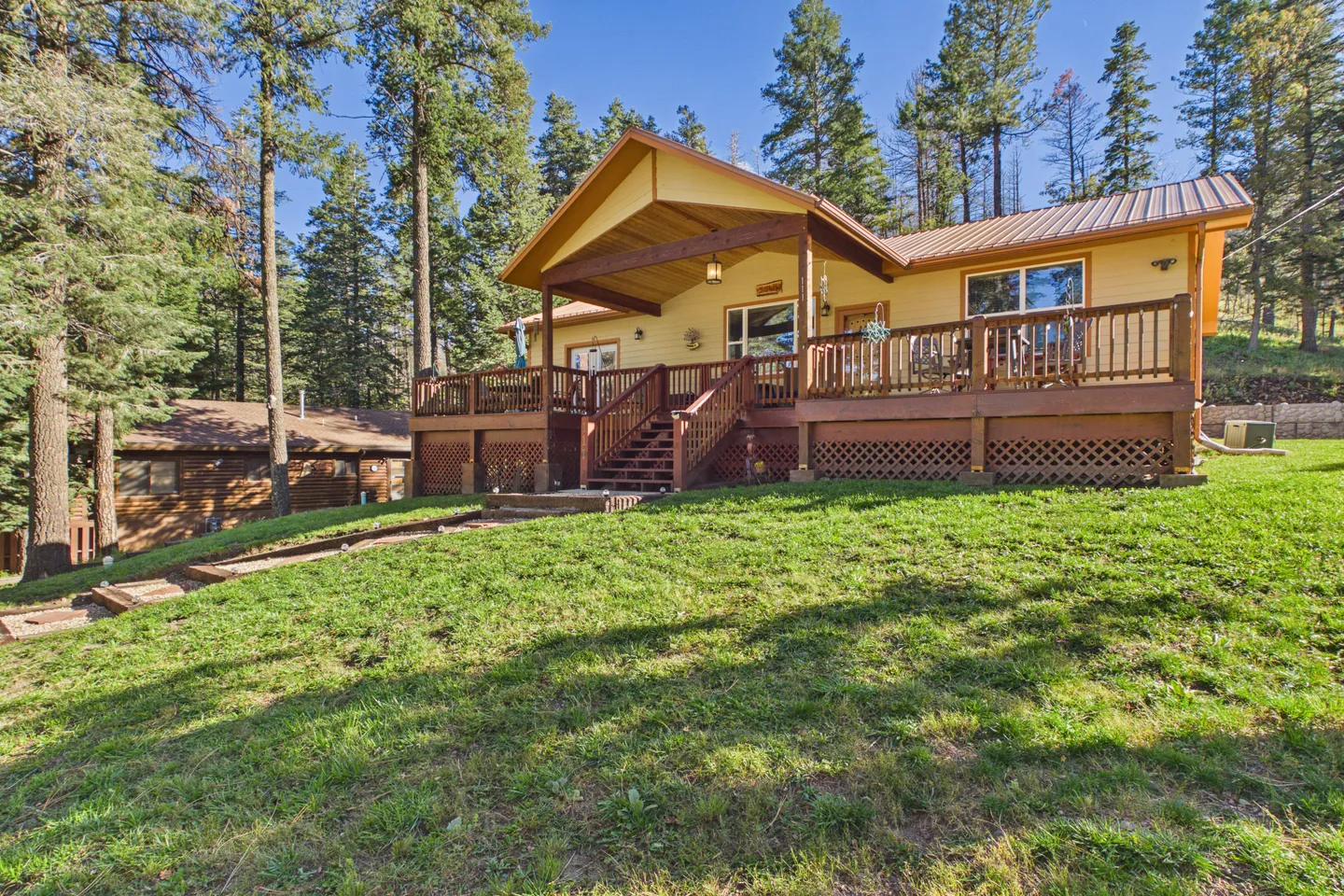 Exterior view of a cabin with a large elevated front deck, surrounded by tall pine trees and a grassy yard.