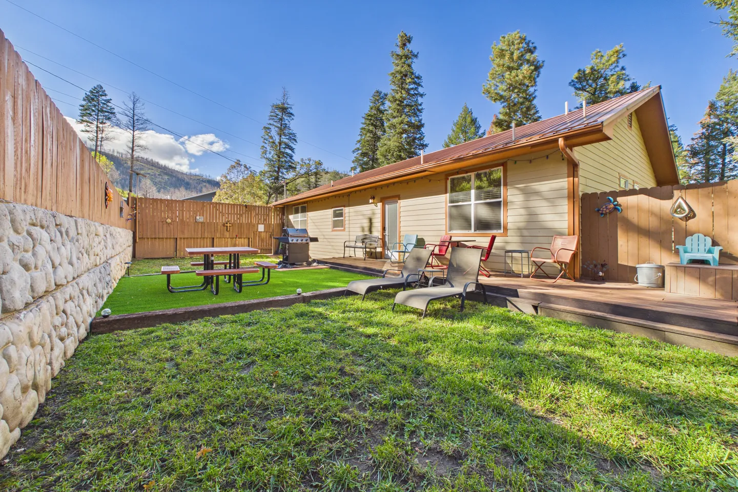 Fenced backyard featuring a green lawn, artificial turf area with a picnic table, barbecue grill, and a raised wooden deck with outdoor seating.