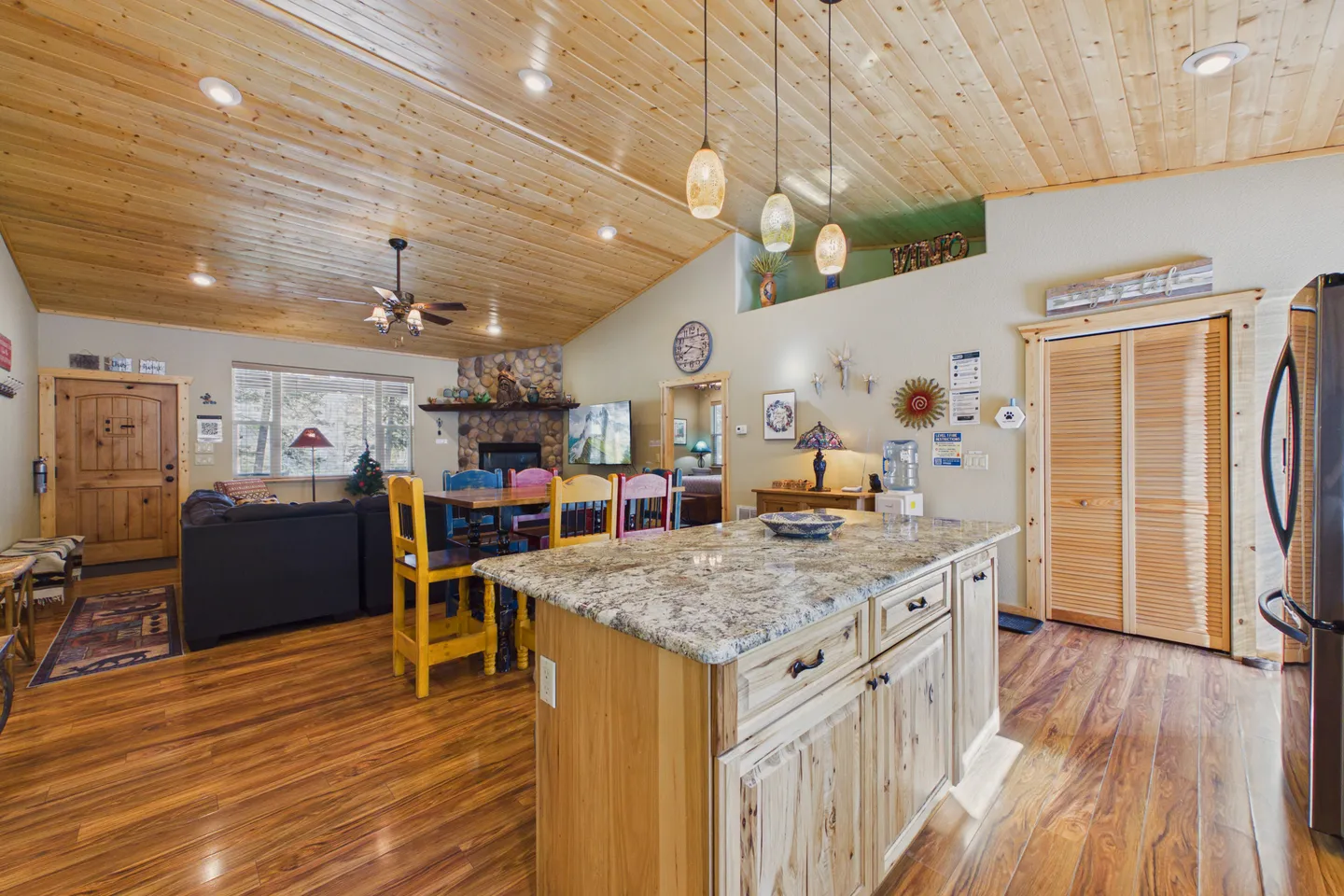 Open-concept living room, dining area, and kitchen featuring vaulted wood-paneled ceilings, a stone fireplace, and a granite-topped kitchen island.