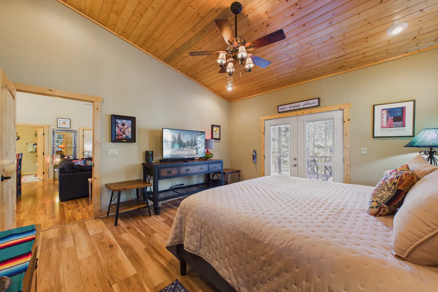 Bedroom with vaulted wood-plank ceiling, wood flooring, a bed, a flat-screen television on a console, and French doors leading to a balcony.