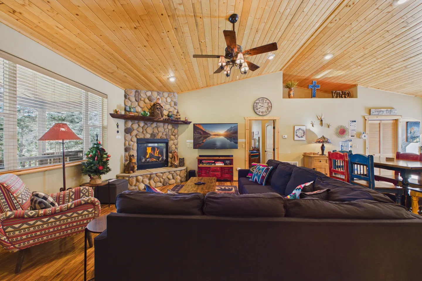 Living room featuring a stone fireplace with a fire burning, wood-plank vaulted ceiling, wall-mounted TV, and dark sectional sofa.
