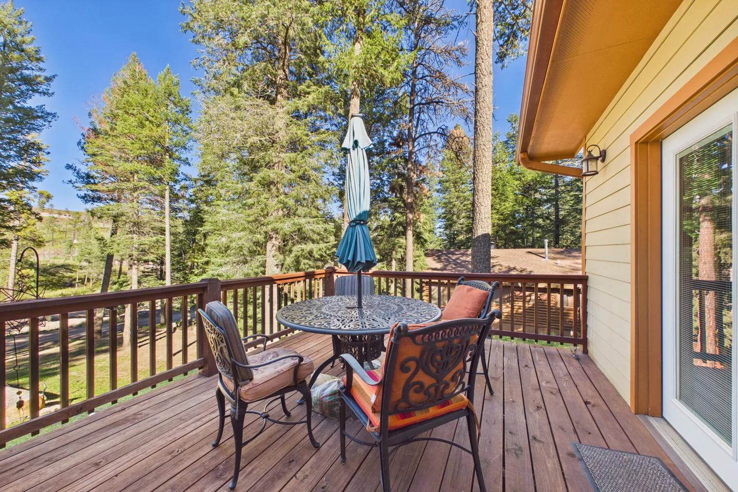 Outdoor wooden deck with a round patio table, umbrella, and chairs overlooking tall pine trees under a blue sky.