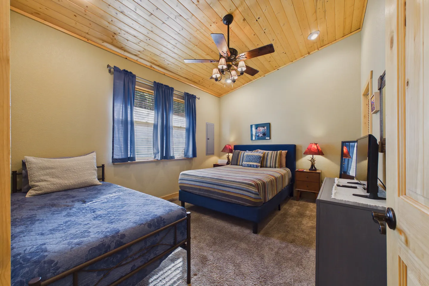 Bedroom with vaulted wood-plank ceiling, ceiling fan, two beds, carpet flooring, and a dresser with a TV.