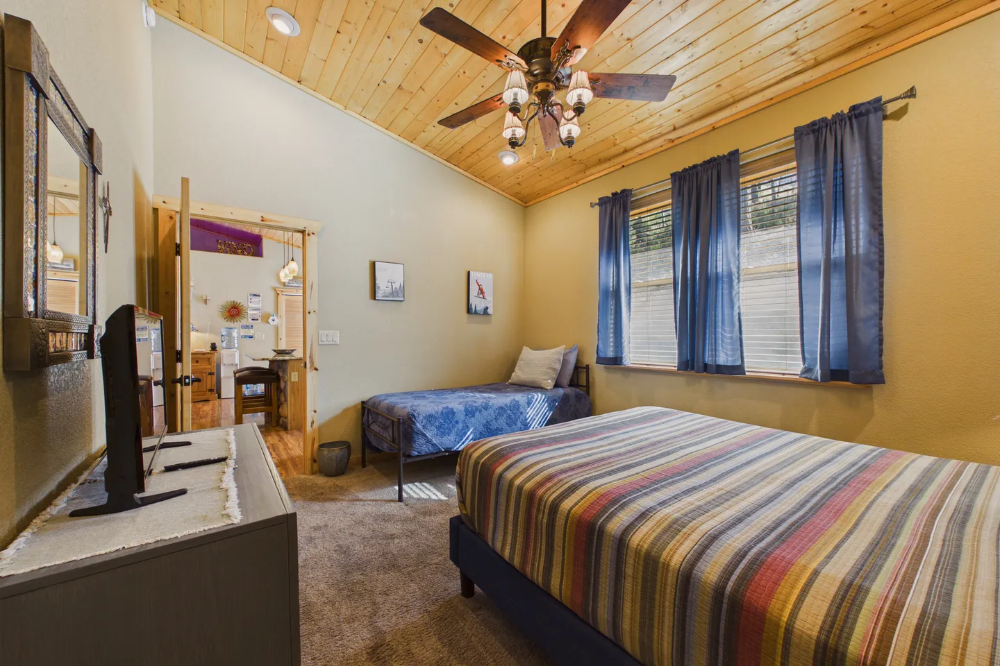 Bedroom with vaulted wood-paneled ceiling, ceiling fan, a larger striped bed, a twin bed, a dresser with a TV, and carpeted flooring.