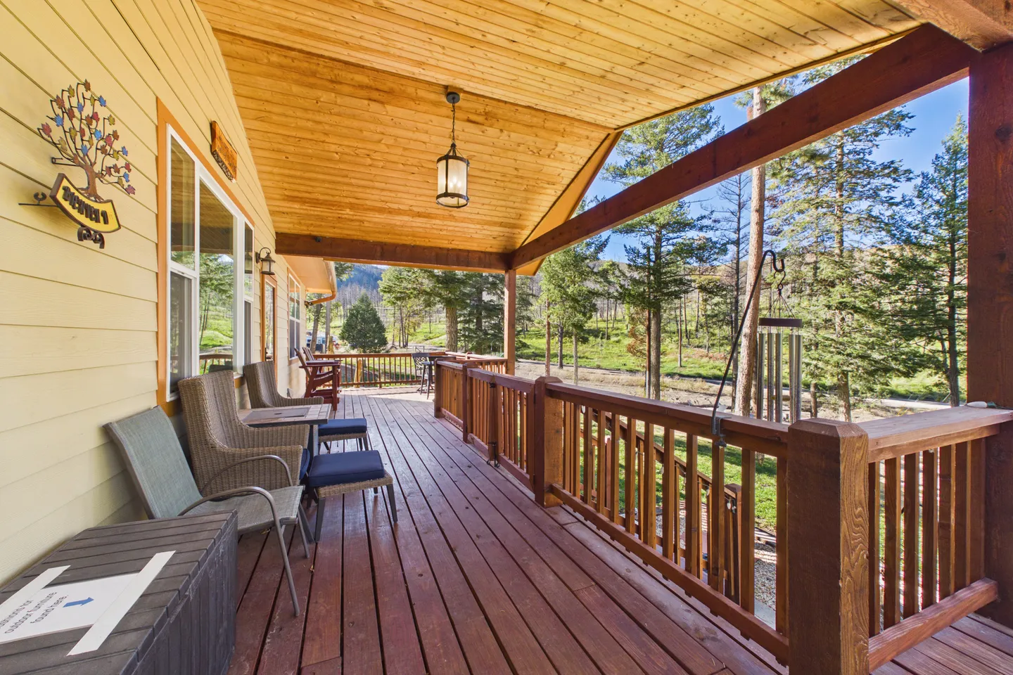A long, covered wooden porch with armchairs, wooden railings, and views of surrounding pine trees on a sunny day.