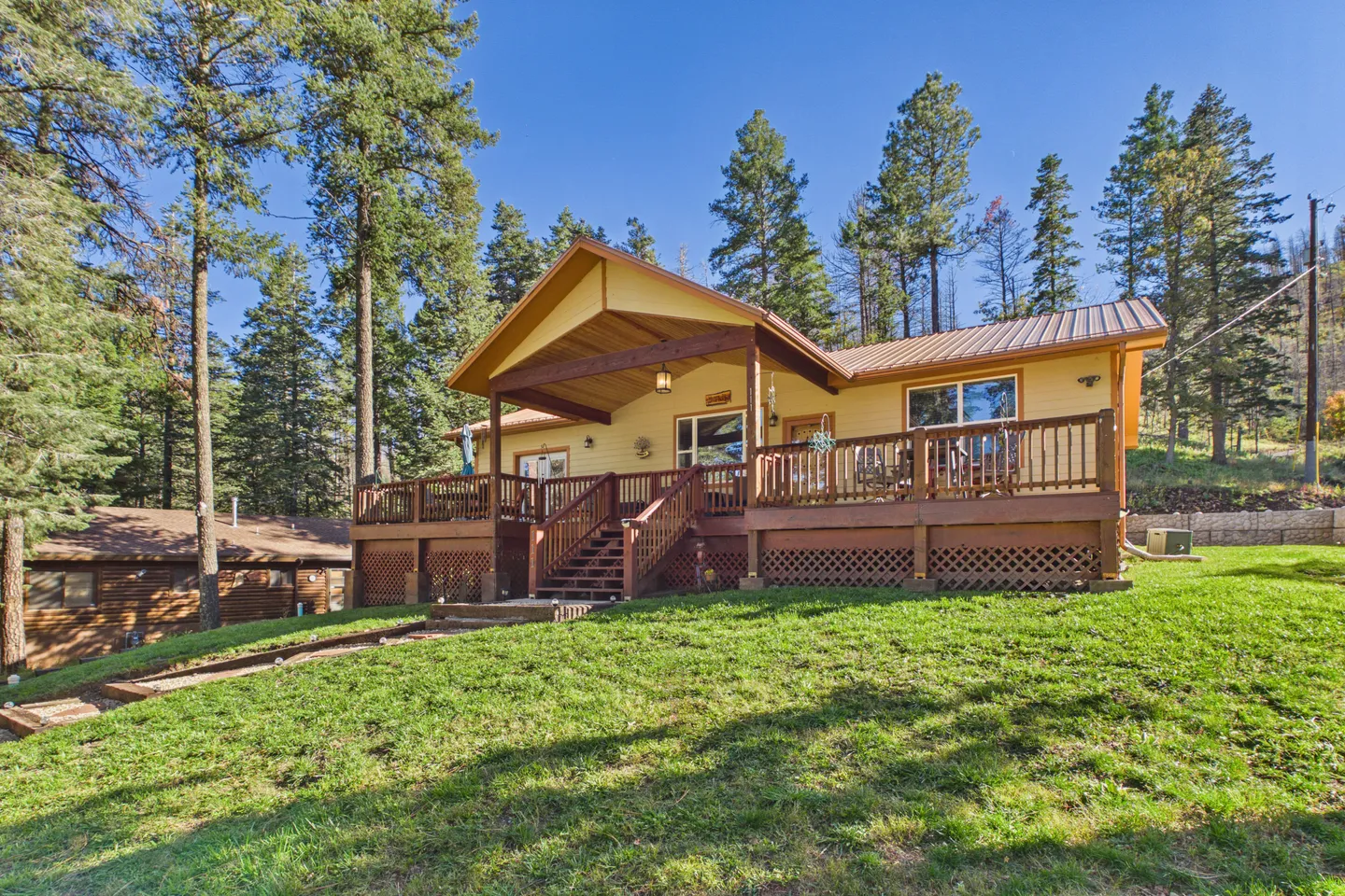 Exterior view of a cabin with a large elevated wooden deck and grass yard surrounded by pine trees.