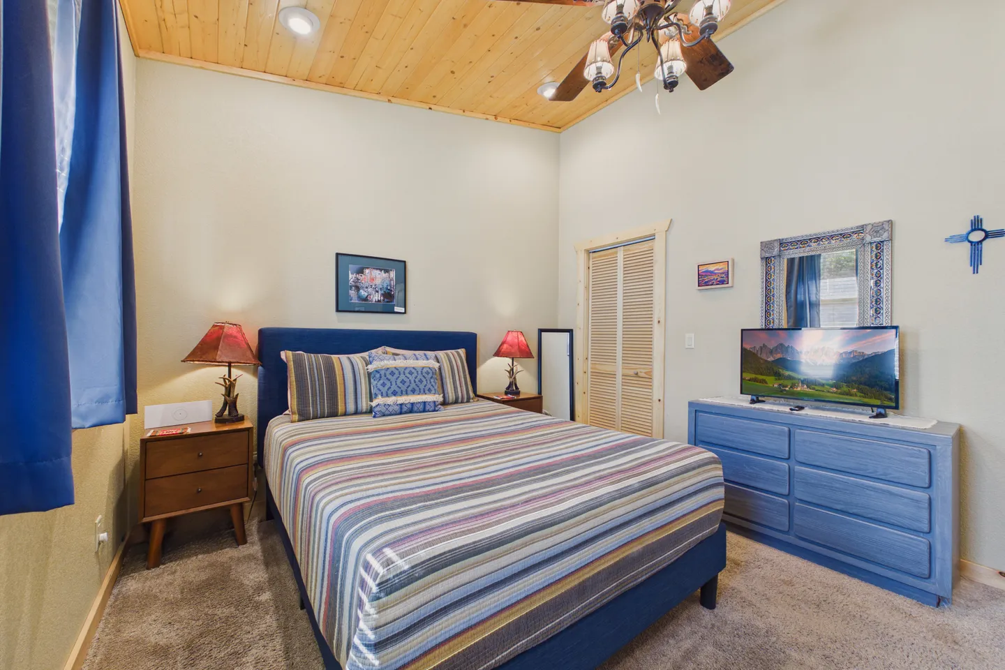 Bedroom with a striped bedspread on a blue bed frame, wood plank ceiling, and a blue dresser with a television.