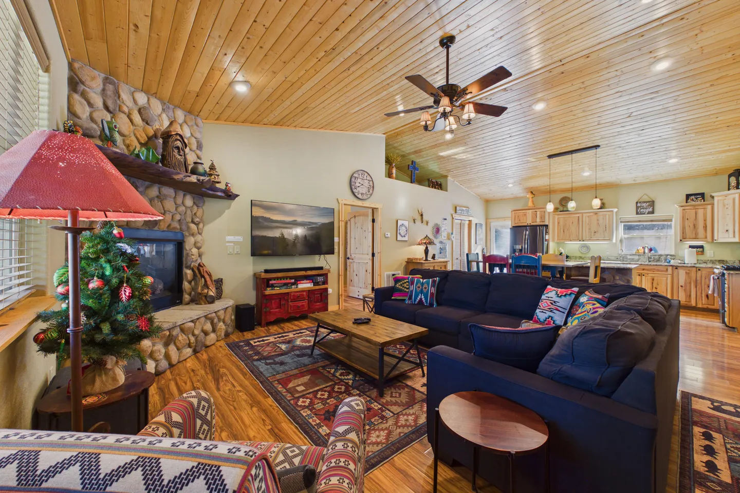 Open-concept living room and kitchen featuring a stone fireplace, wood-paneled vaulted ceiling, and dark sectional sofa.
